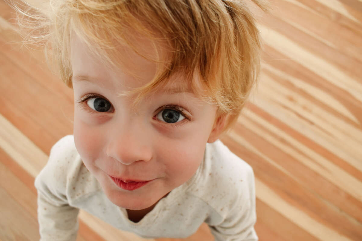 A 4 year old boy with blonde hair and blue eyes wearing a cream jumper looks up to the camera and has a whimsical expression on this face.