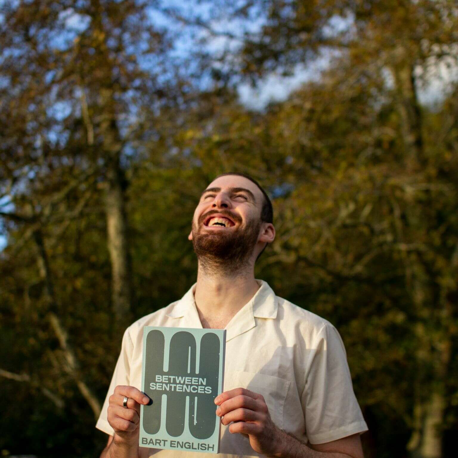 Bart has short, cropped hair and a beard, he looks up smiling holding his book cover. He's in front of some trees