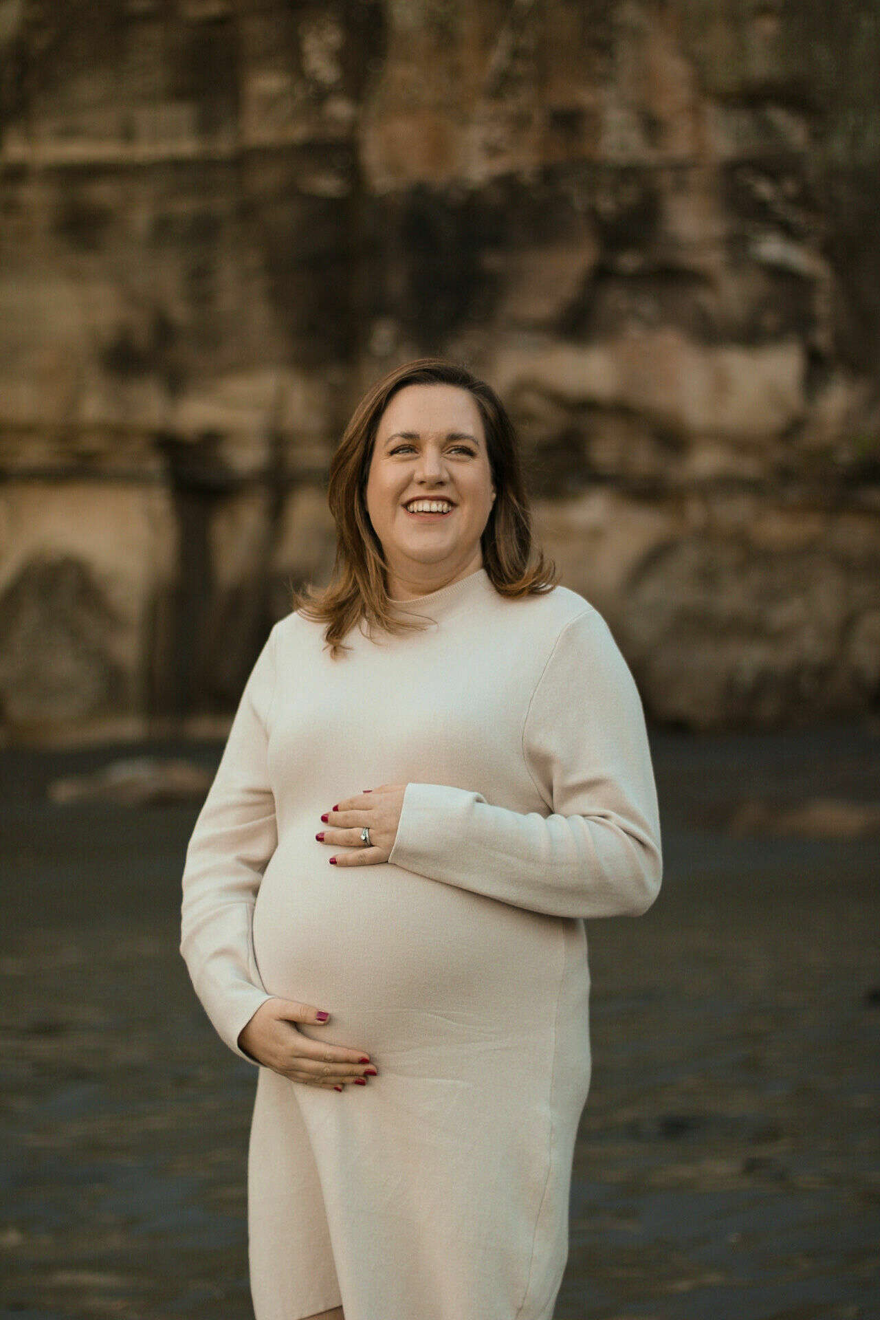 Image shows Natalie standing on a beach wearing a white long sleeve dress holding her baby bump laughing towards the camera