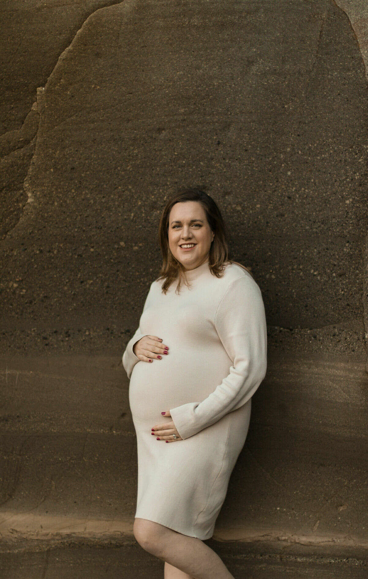 Natalie holds her pregnant belly wearing a cream jumper dress and stands against a cliff rock face. She has brown shoulder length hair and is smiling into the camera
