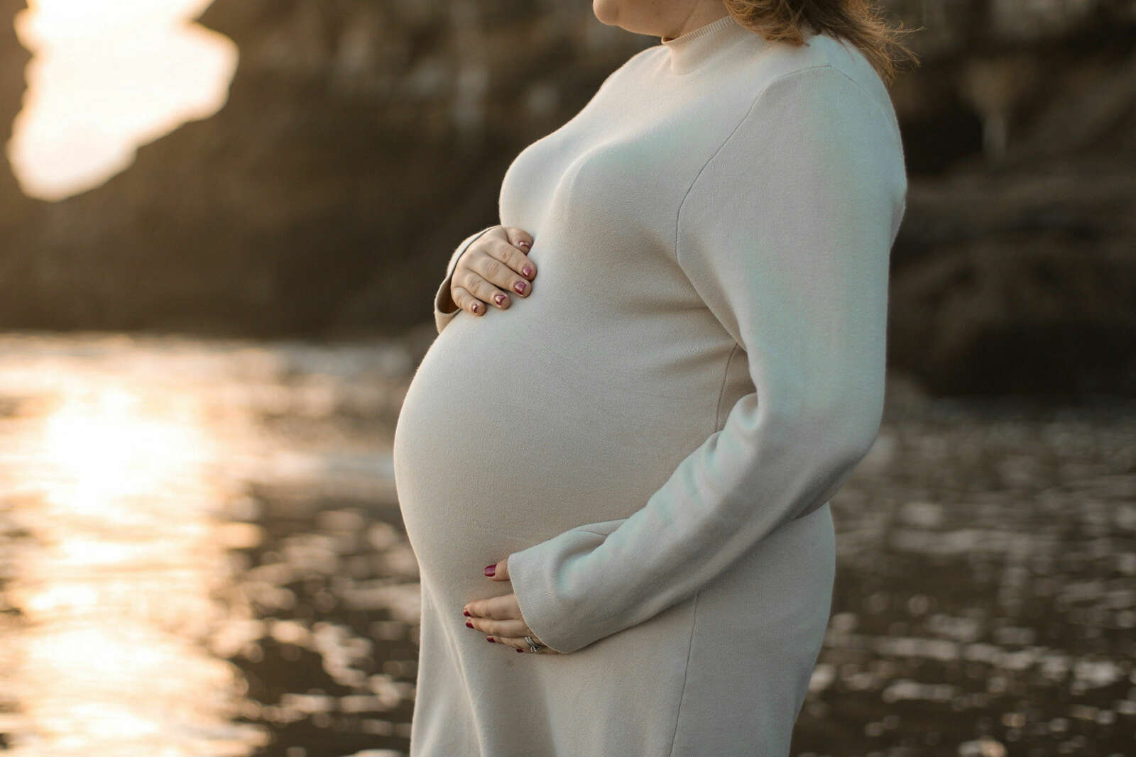 Image shows Natalie standing in a beach wearing a white long sleeve dress holding her baby bump looking towards the sunset.