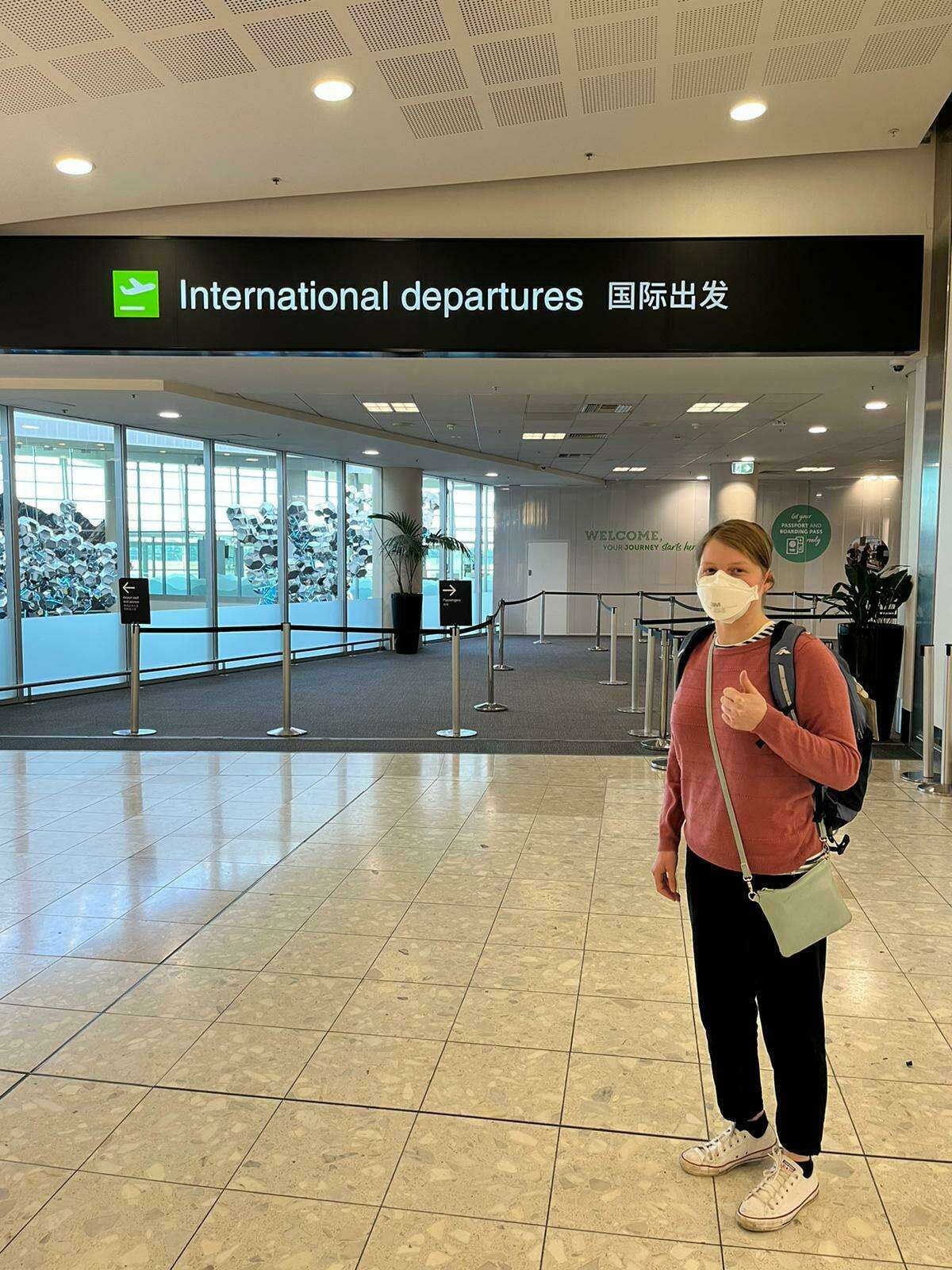 Image shows Victoria standing in an airport under the sign 'International departures'. She is wearing a red top, black pants and sneakers with a mask. Her hair is pulled back and she is carrying a backpack as she looks towards the camera.