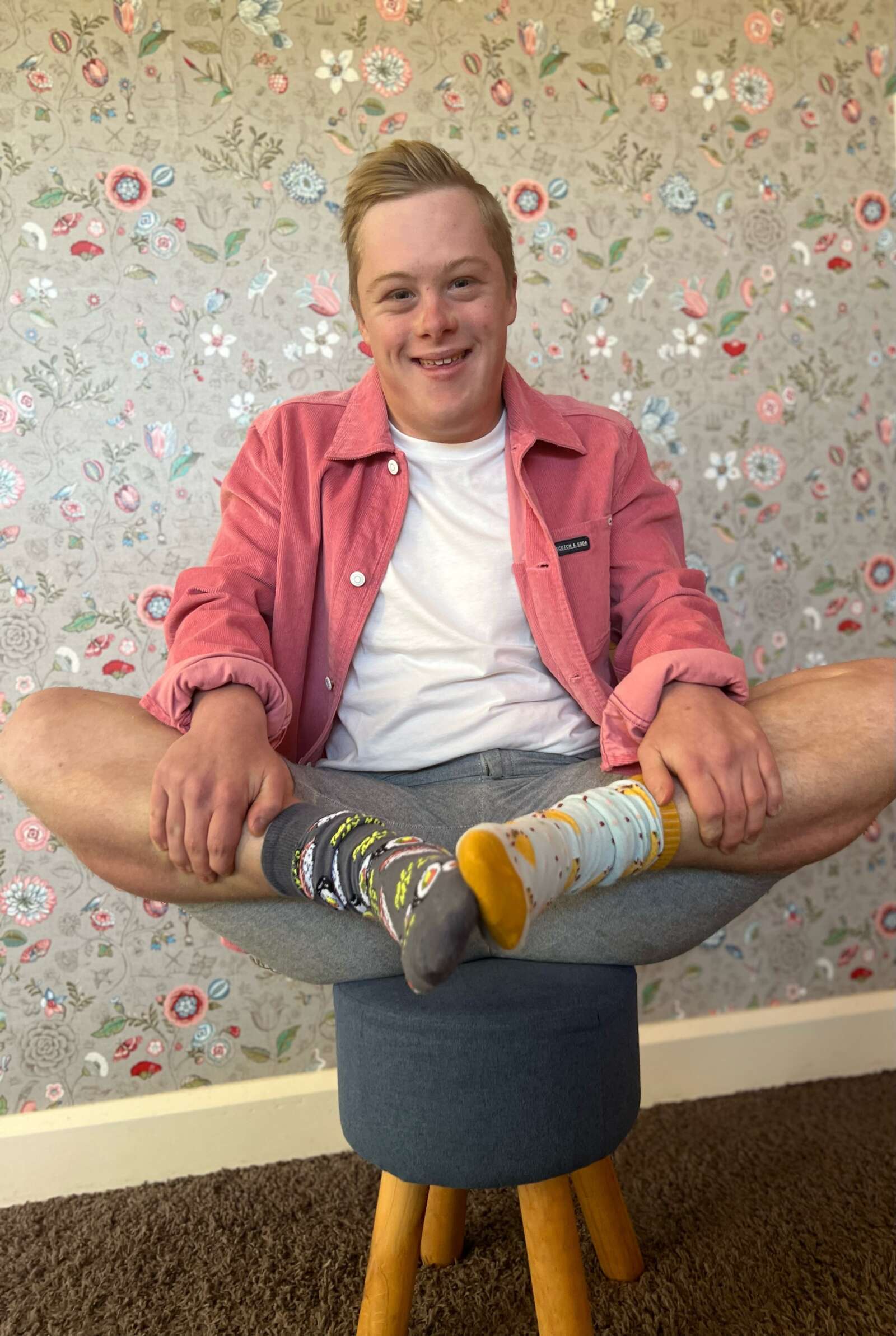 Luka sits on a stool in a red shirt, white tee and grey shorts. He is holding his feet up showing off his odd socks in recognition of World Down syndrome Day.