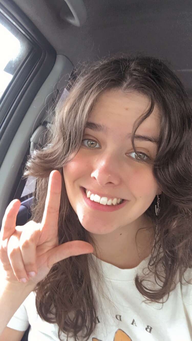 A photo of Hope Cotton looking into the camera doing the NZ sign language sign for 'I love you'. She is wearing a white t-shirt and has long brown curly hair.