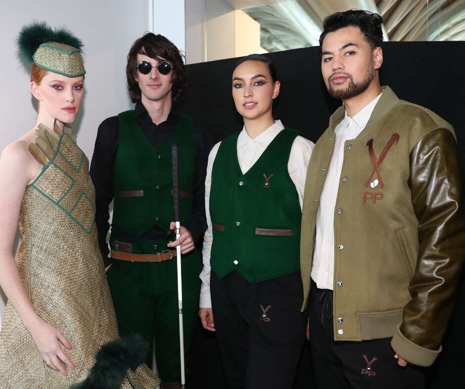 Ari is backstage with 3 other models for the Susana Task show. From left to right. Model wears a beige and green dress and hat, she has read hair and matching makeup. Ari stands next to her and is wearing a green waistcoat and shorts, with a black shirt and brown belt holding his cane. The model on the other side of him wears a white shirt, green waistcoat and black pants and has her hair tied back. The model next to her wears a white tee, and greener brown jacket with black pants. He has short dark hair and facial hair.