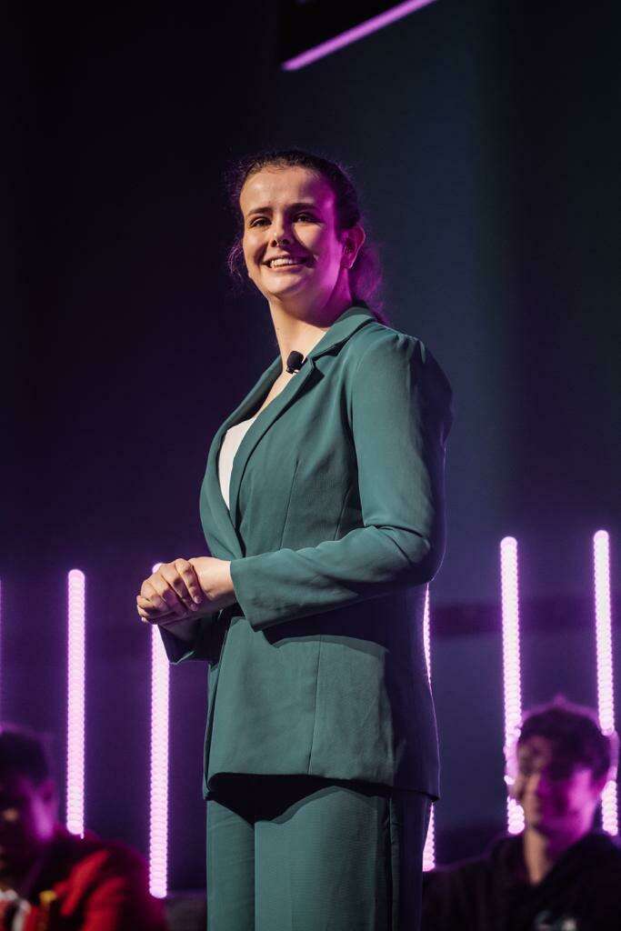 Hope Cotton giving a speech at an event in Auckland. She is standing on stage wearing a green suit and white top with her hair pulled back in a ponytail. She has a backdrop of pink neon lighting behind her.