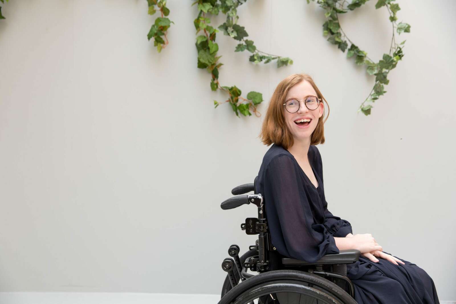 Woman wearing glasses laughs in navy dress, in front of green foliage. she is using a wheelchair