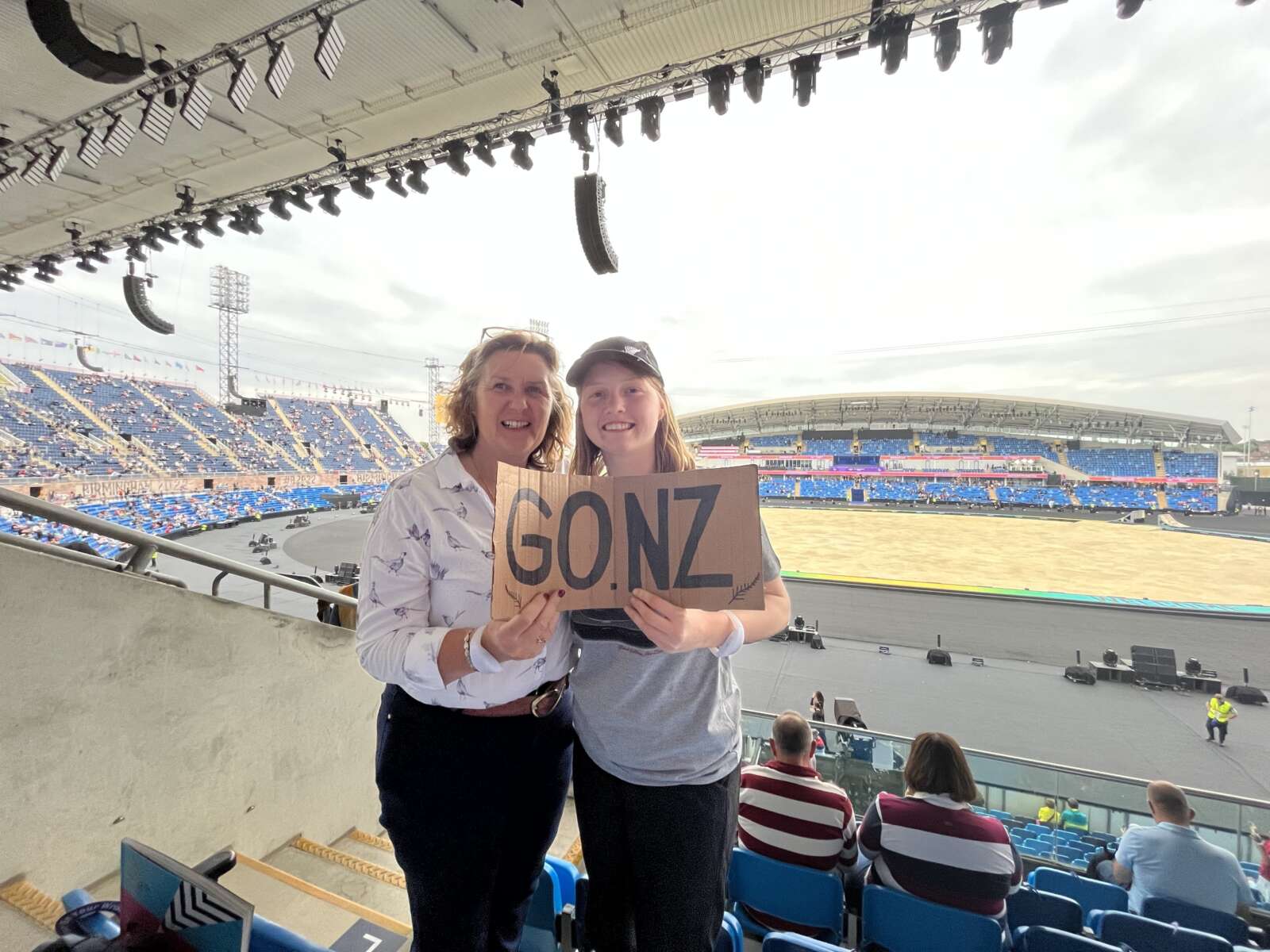 Victoria attending the Commonwealth games with a family member. Both are standing inside the stadium looking at the camera with the arena behind them holding a sign saying 'Go NZ'