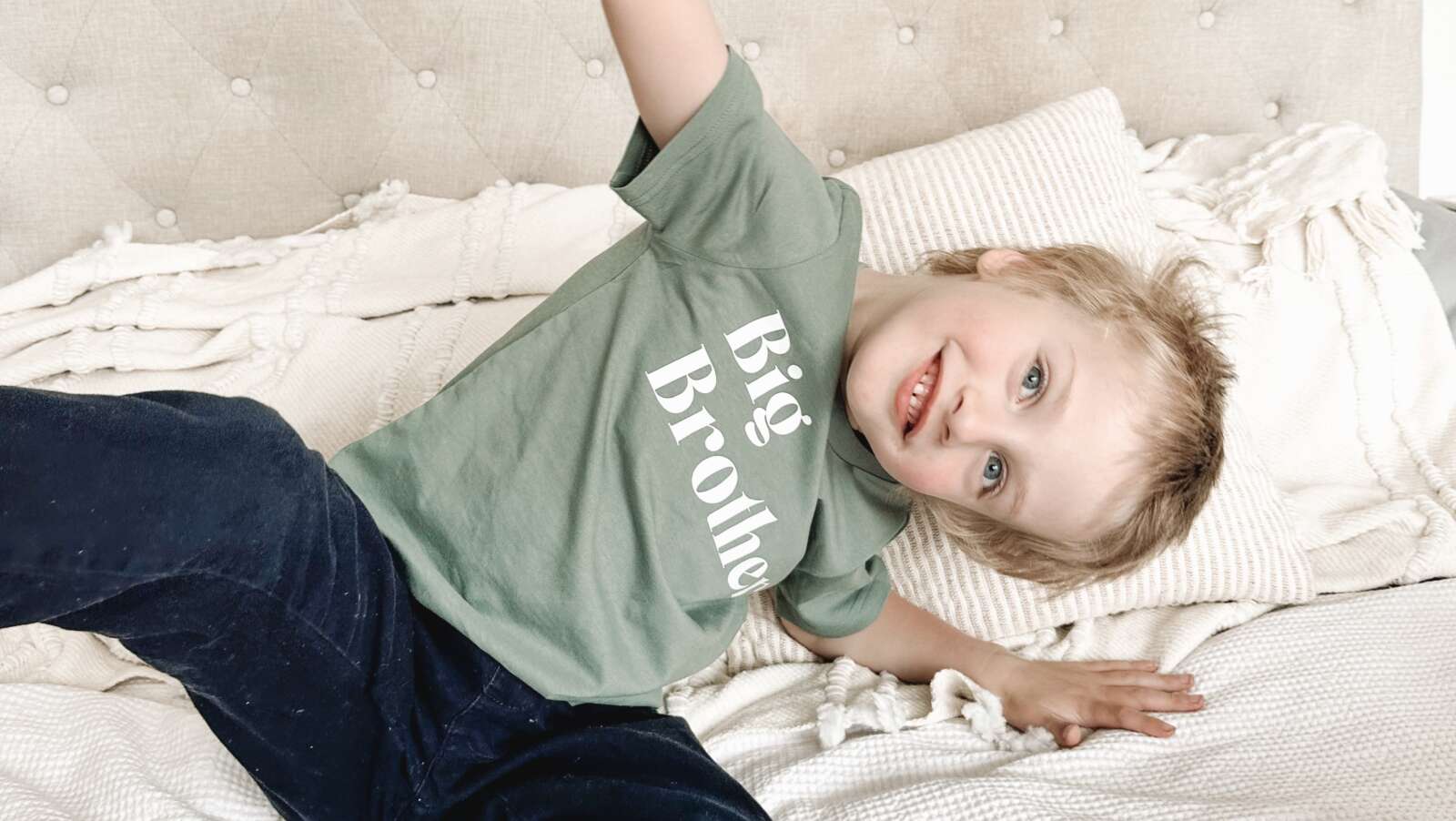 A preschooler playfully sits on a bed wearing navy trousers and a green tee shirt. His head is turned to the side, with his blue eyes looking into the camera.