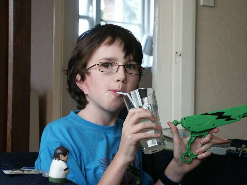 An image of Zach as a child, he has brown hair, wear glasses and is drinking from a cup with a straw as he looks into the camera. he is in a home wearing a blue t-shirt.