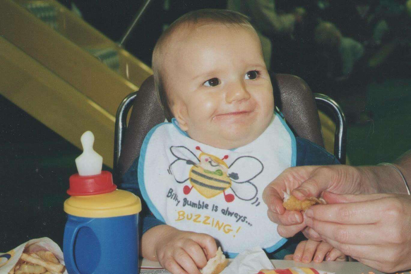 Image shows Zach when he was a baby. He is sitting in a high chair wearing a bib with a bee on it and looks up at an adult.