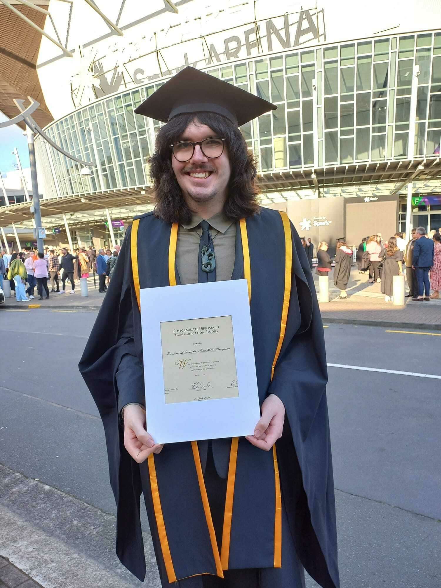 Photo shows Zach graduating as he stands in front of Spark Arena in Auckland. he is wearing his graduation robes and holds his certificate proudly smiling at the camera. He has long wavy brown hair and glasses.