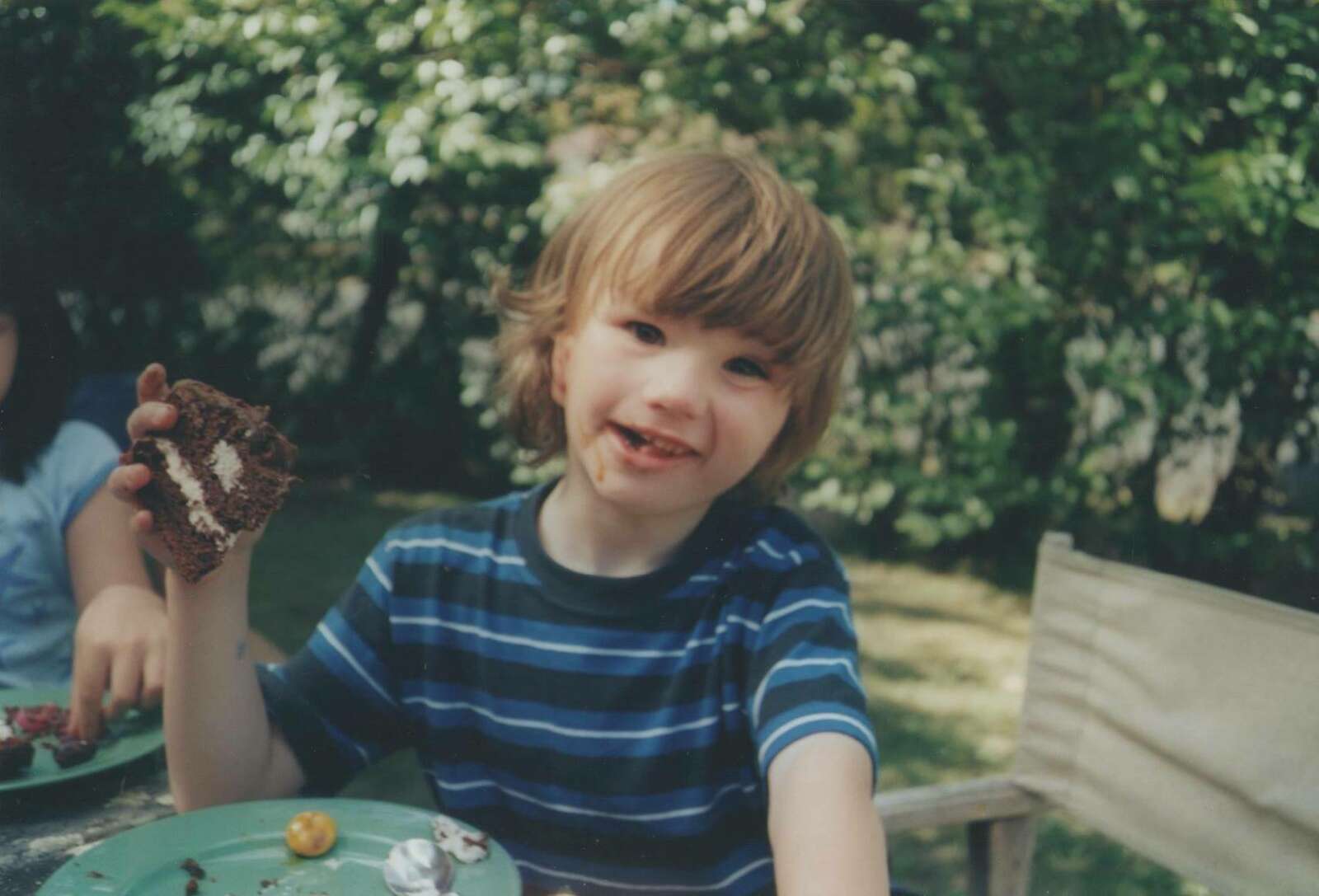 Photo of Zach when he was a child. The image shows a young Zach with brown hair, brown eyes and wearing a blue striped tshirt looking at the camera while holding a slice of chocolate cake as he sits in a garden.