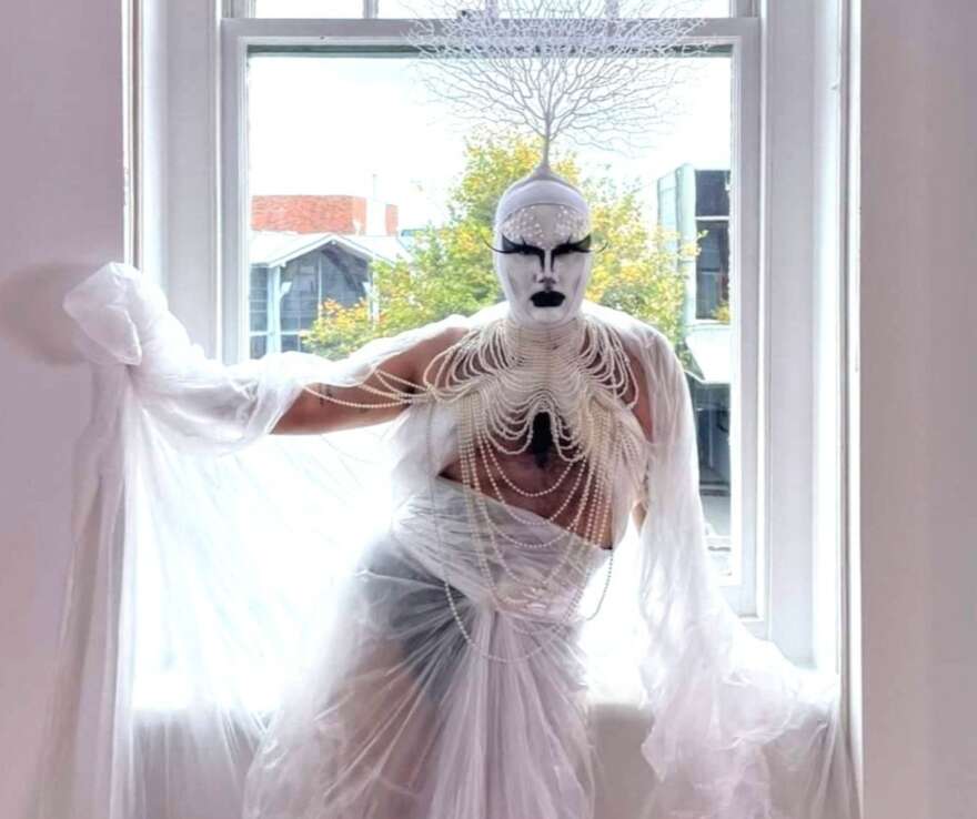 Image shows Misty leaning against a window in a stage costume. The costume is made of white sheer fabric and is draped over them. They are wearing black underwear and a white head piece with black eye makeup and lips.