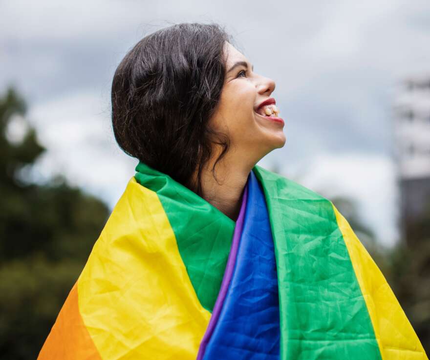 Mamie looks away from camera with pride flag over shoulder. Long brown curly hair.