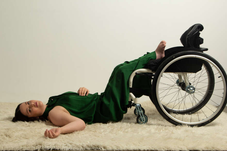 Bec is wearing a green jumpsuit with her legs up on her wheelchair as she lays on the ground on a white fluffy rug. She has shoulder length dark hair.
