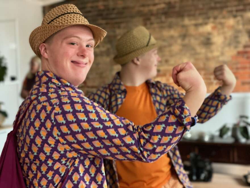 Luka has a hat on and smiles looking at the camera as he flexes his muscles. He is wearing a straw yellow hat, white t-shirt and patterned orange shirt.