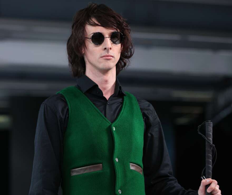 Ari on the end of NZFW runway, wearing a green waistcoat, black shirt and has his cane his your hand. He has ear length black hair and wears sunglasses.