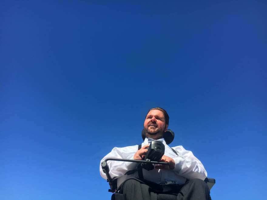 Man in an electric wheelchair wears a white shirt and looks out in front of a blue sky