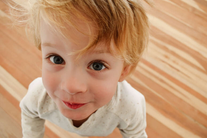 A 4 year old boy with blonde hair and blue eyes wearing a cream jumper looks up to the camera and has a whimsical expression on this face.