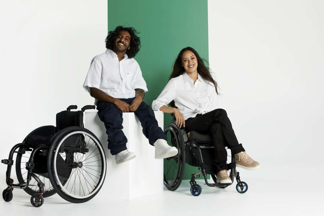 Shakti and chelsea sit near their wheelchairs on a set, in front of a green wall.