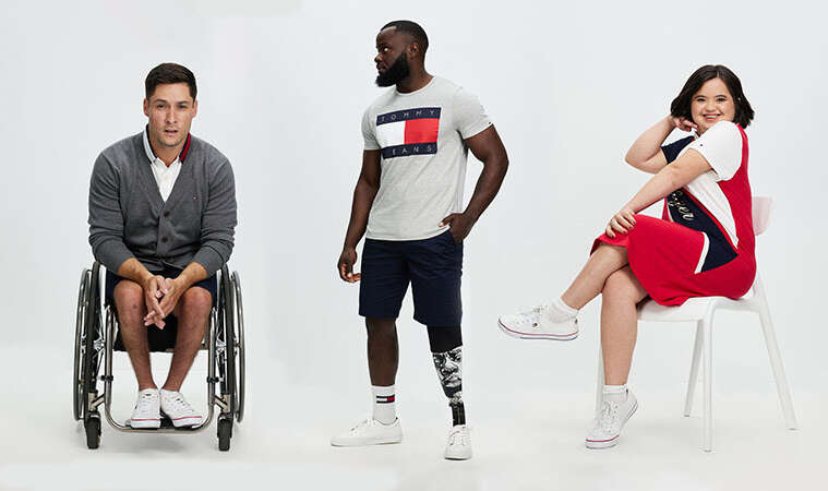 A group of disabled models lined up next to one another. From left man sits in wheelchair with his hands in lap, he wears a grey sweatshirt and white shoes. In middle, a man stands tall and to the left, he has one artificial limb you can see through shorts and a grey t-shirt, the last woman lives with Down Syndrome, she sits on a chair with a red and navy and white dress on.