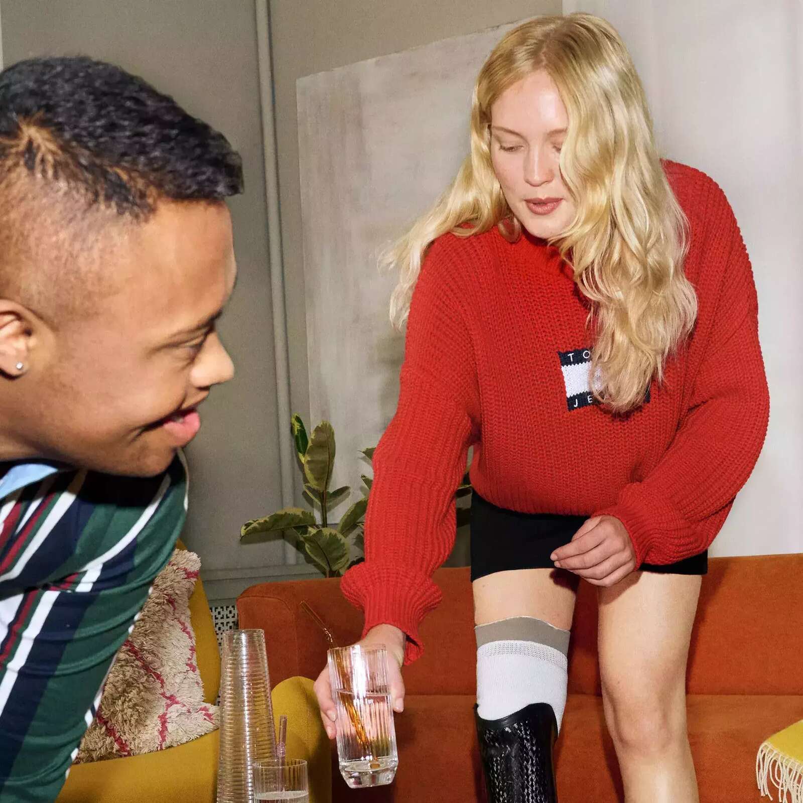 Image is of a blonde amputee leaning down putting a class on a table, a man with Down Syndrome is closer in the frame, he has brown hair and features. The two are hanging out together.