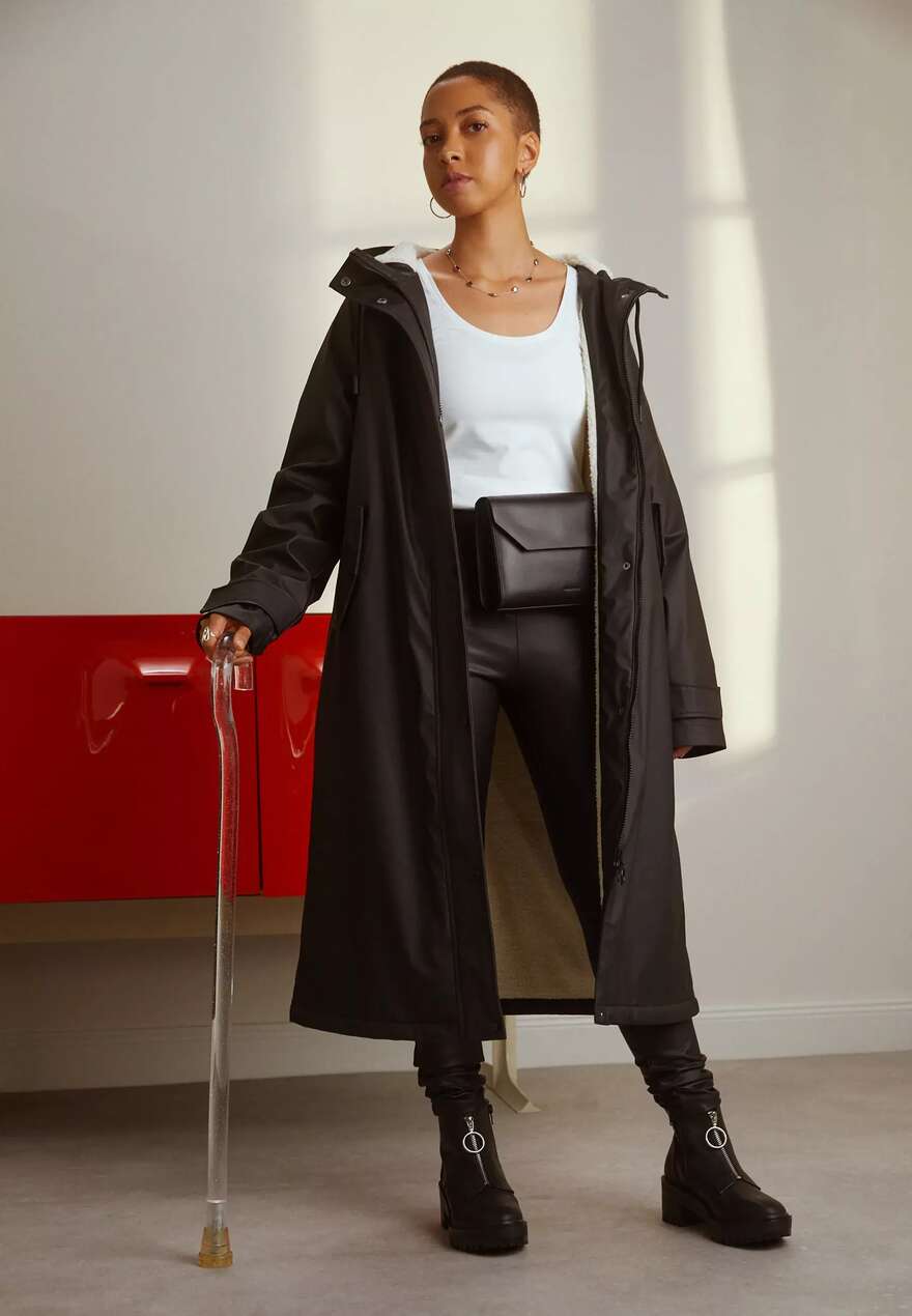Image is of a person with tan skin and brown hair, they have a buzz cut and use a cane. They wear a brown coat, a white tank top and leather trousers.