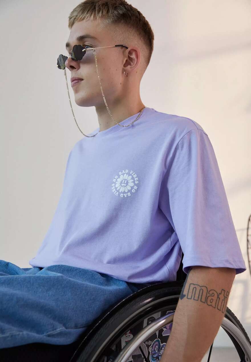 Image is of a wheelchair user, looking away from camera. He has blonde hair, fair features and wears purple sunglasses with an adaptive purple tee shirt