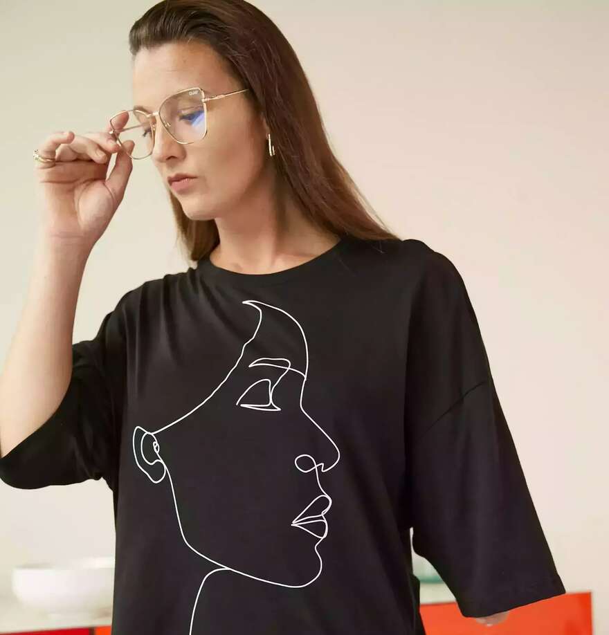 Image is of a woman with fair skin and brown hair, she wears clear rimmed glasses and is an upper limb amputee, her shirt has the outline of a womans face on it.