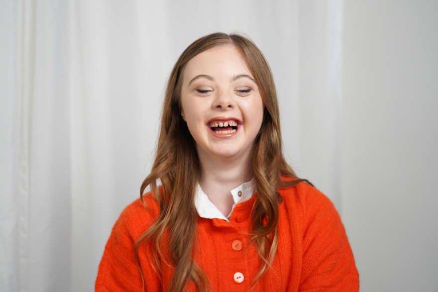 Mid laugh, Libby who lives with Down Syndrome is photographed. She has curled hair, an orange jumper and red lipstick on.