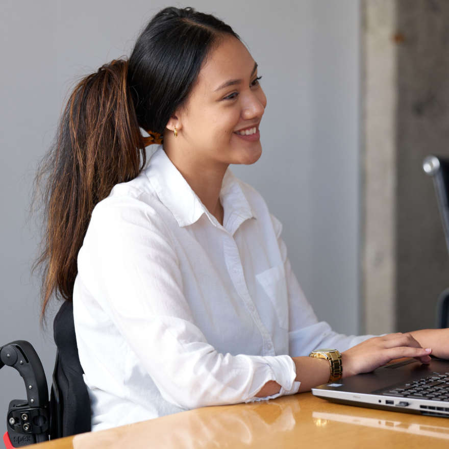 Chelsea wears a white shirt, and sits at a table in her wheelchair as she types on a computer.