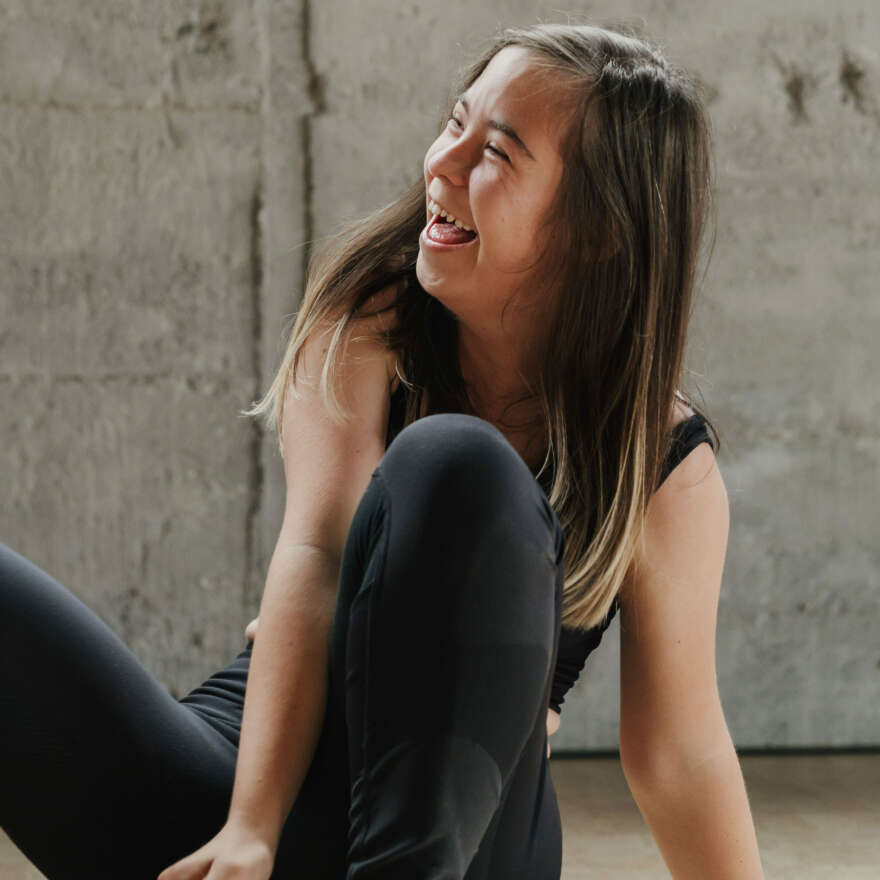 Molly wears athletic gear and smiles she has brown hair and is in front of a concrete dance studio wall.