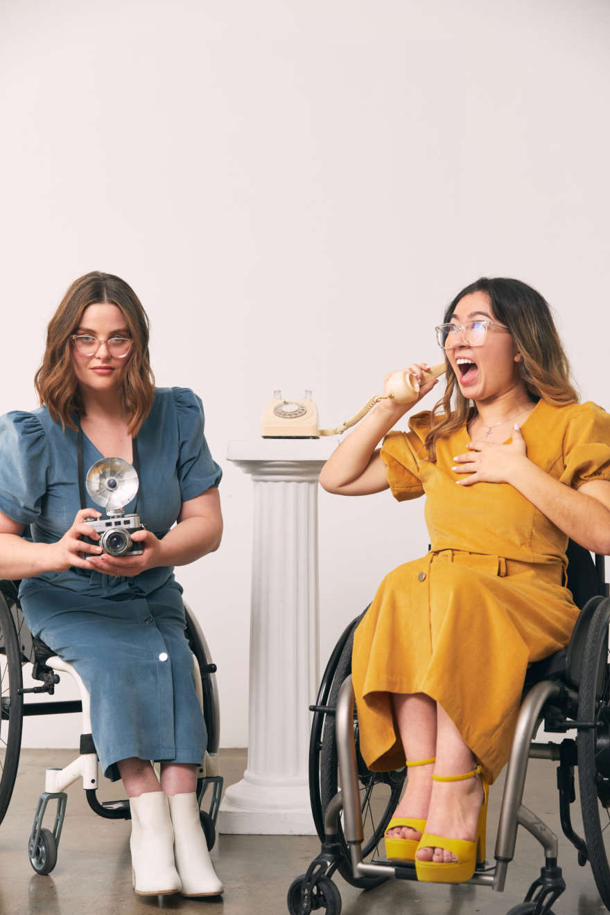 Two models, wheelchair users in retro mustard and deep blue outfits, are answering the phone, one holds a camera. They are making old school journalist faces.