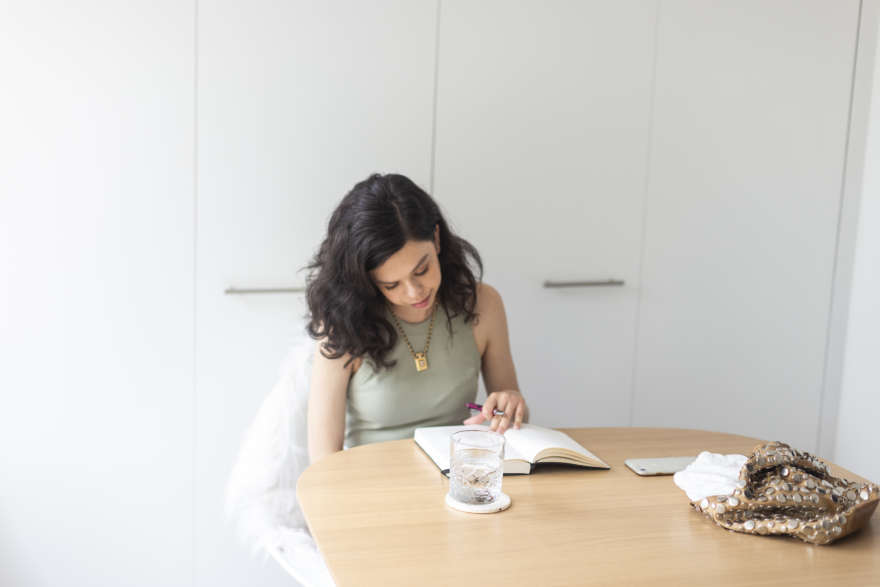 A young woman with pixie like features, is writing in her journal in a large room against a white wall.