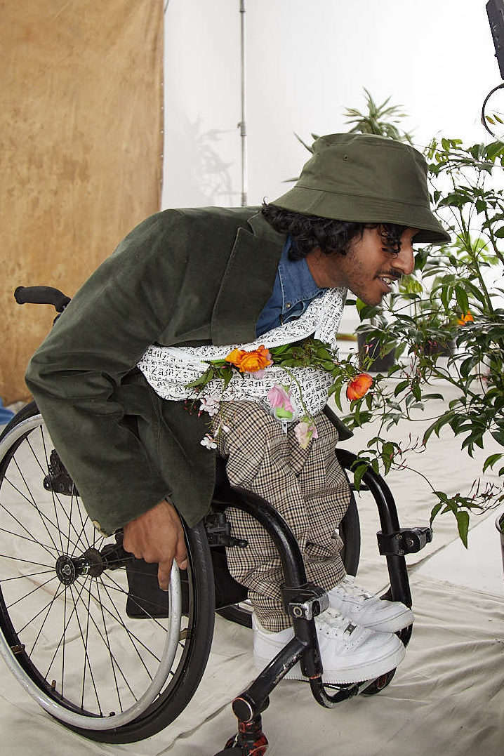 Shakti, a wheelchair user has a hat, trousers and sneakers on as well as a bum bag full of flowers. He's on a set and is kneeling down to the camera's level.