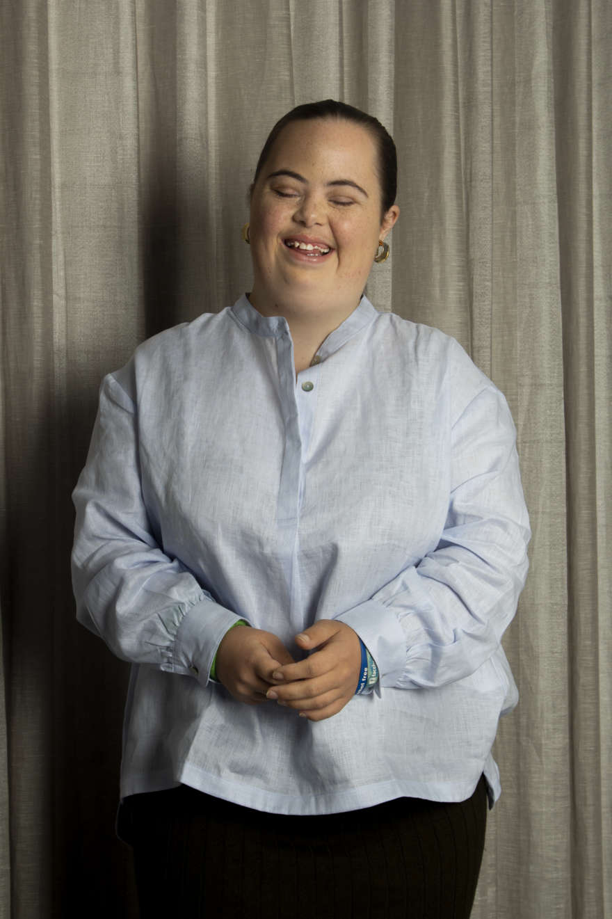 Emma is in a blue linen shirt, standing against a dark grey curtain, she is mid laugh and has her hands together.