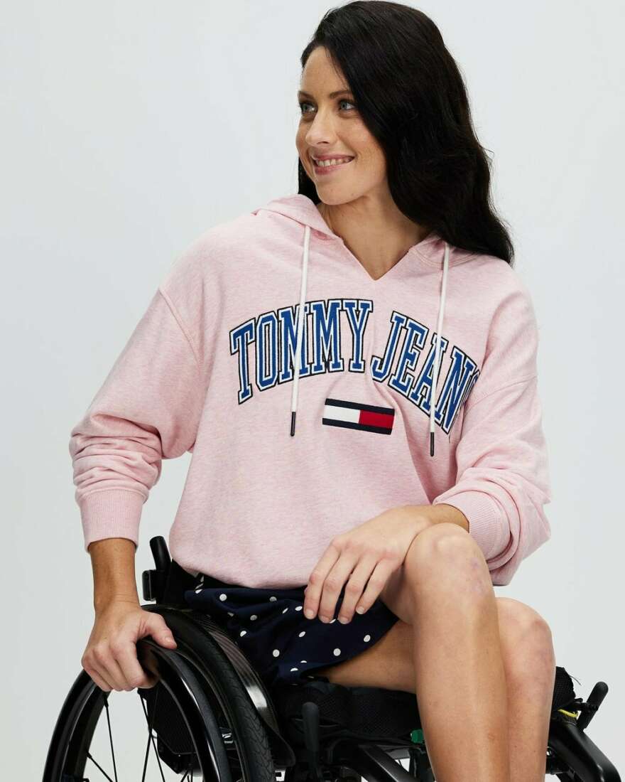 Lauren sits in her wheelchair, she is side on with her legs crossed. She wears a pink jersey with a polka dot dress underneath.