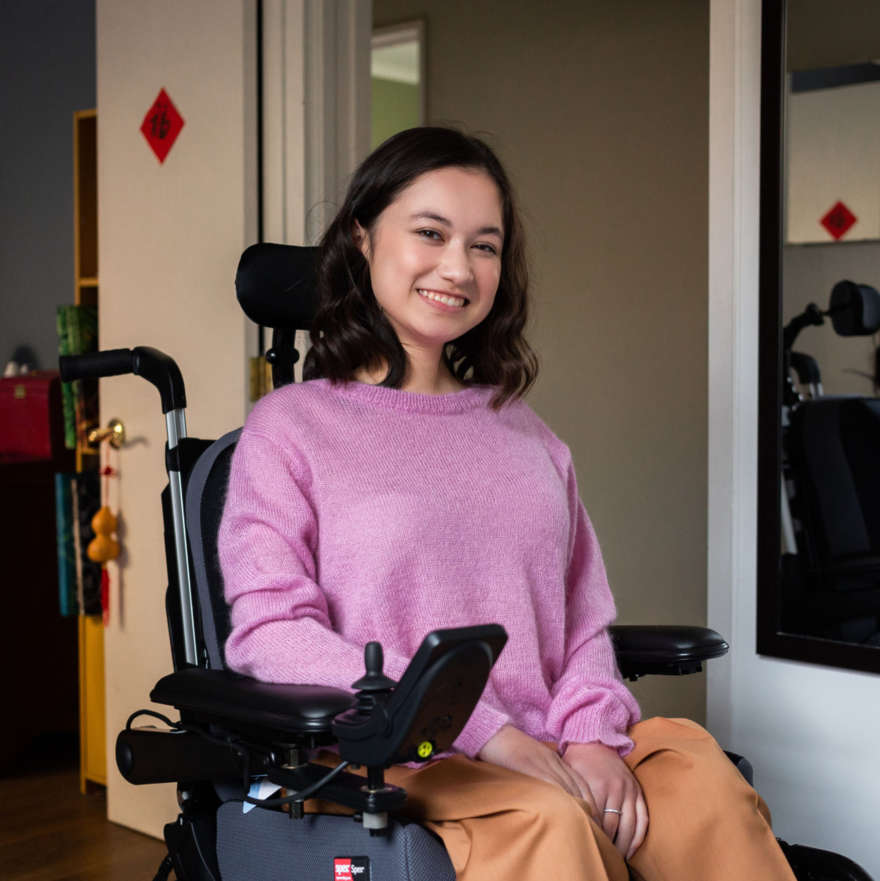 Kelsey is a wheelchair user, she's looking at the camera and smiling, she wears a pink jersey and tan pants.