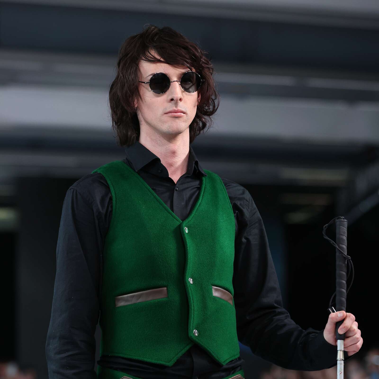Ari is a fair-skinned male with long brunette wavy hair. He is wearing black circular glasses and a black long-sleeve shirt with a green vest. He is walking with a cane.