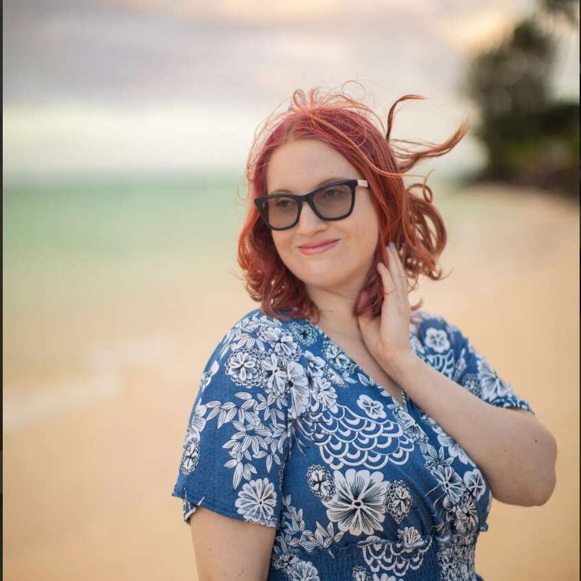 Natasha has dark red hair and fair-skin. She is standing at the beach wearing black sunglasses and a blue dress with white patterns.