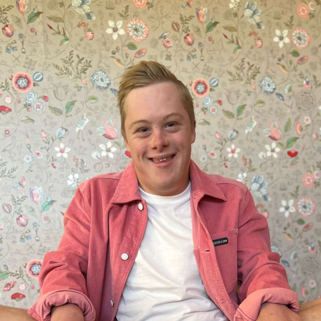 Luka is smiling at the camera. He has short blonde hair. He is wearing a white tee with a red shirt overtop in front of a floral wall.