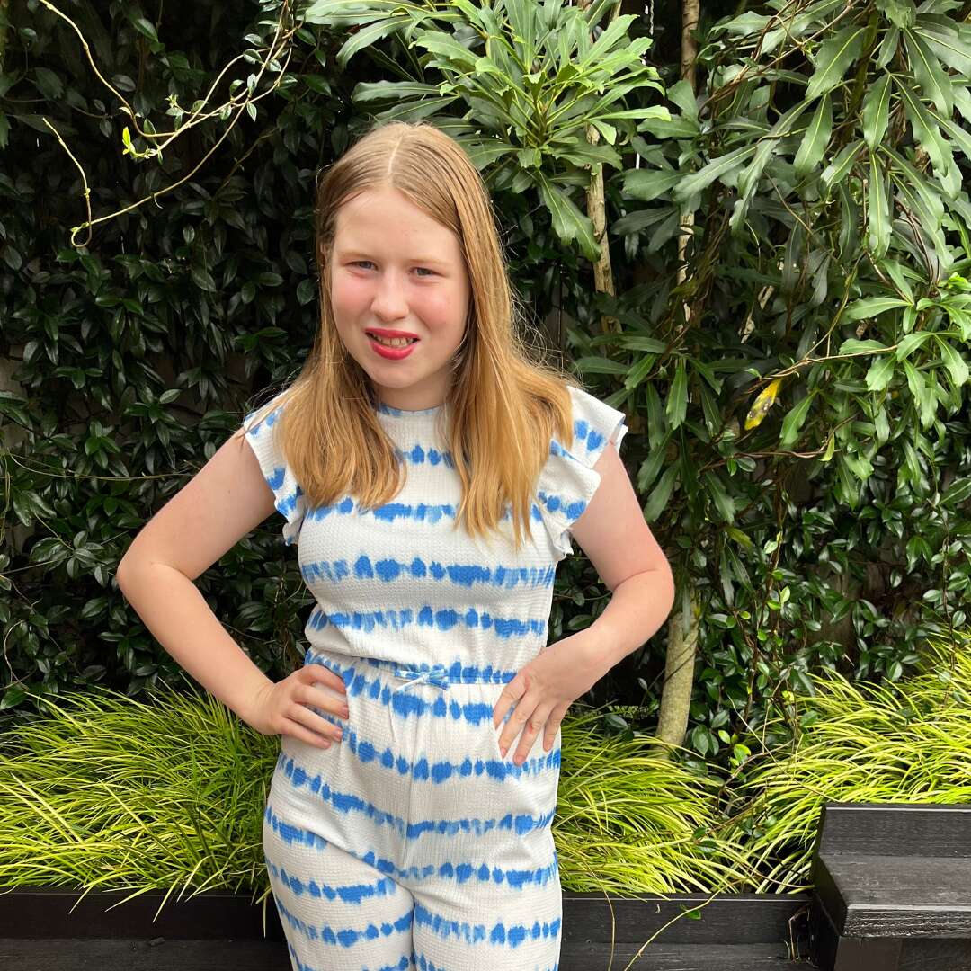 Molly has light hair and is wearing a white and blue striped dress. She is standing in front of a garden