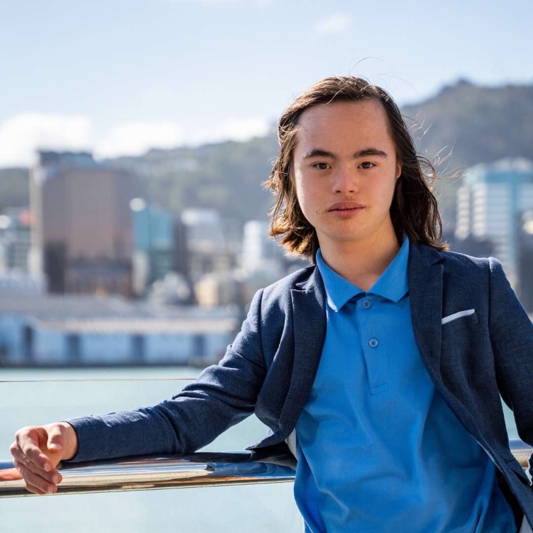 Xavier is an asian male with long black hair. He is standing in a blue shirt and navy blazer. He is standing with the city in the background.