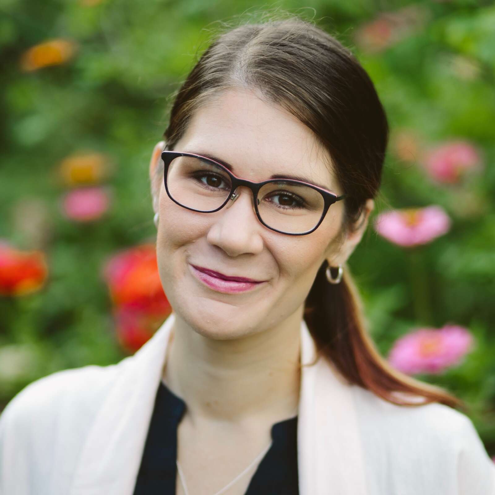 Kylee is a brown-haired female with glasses and hair in a ponytail. She is wearing a light pink blazer with an out of focus floral background.