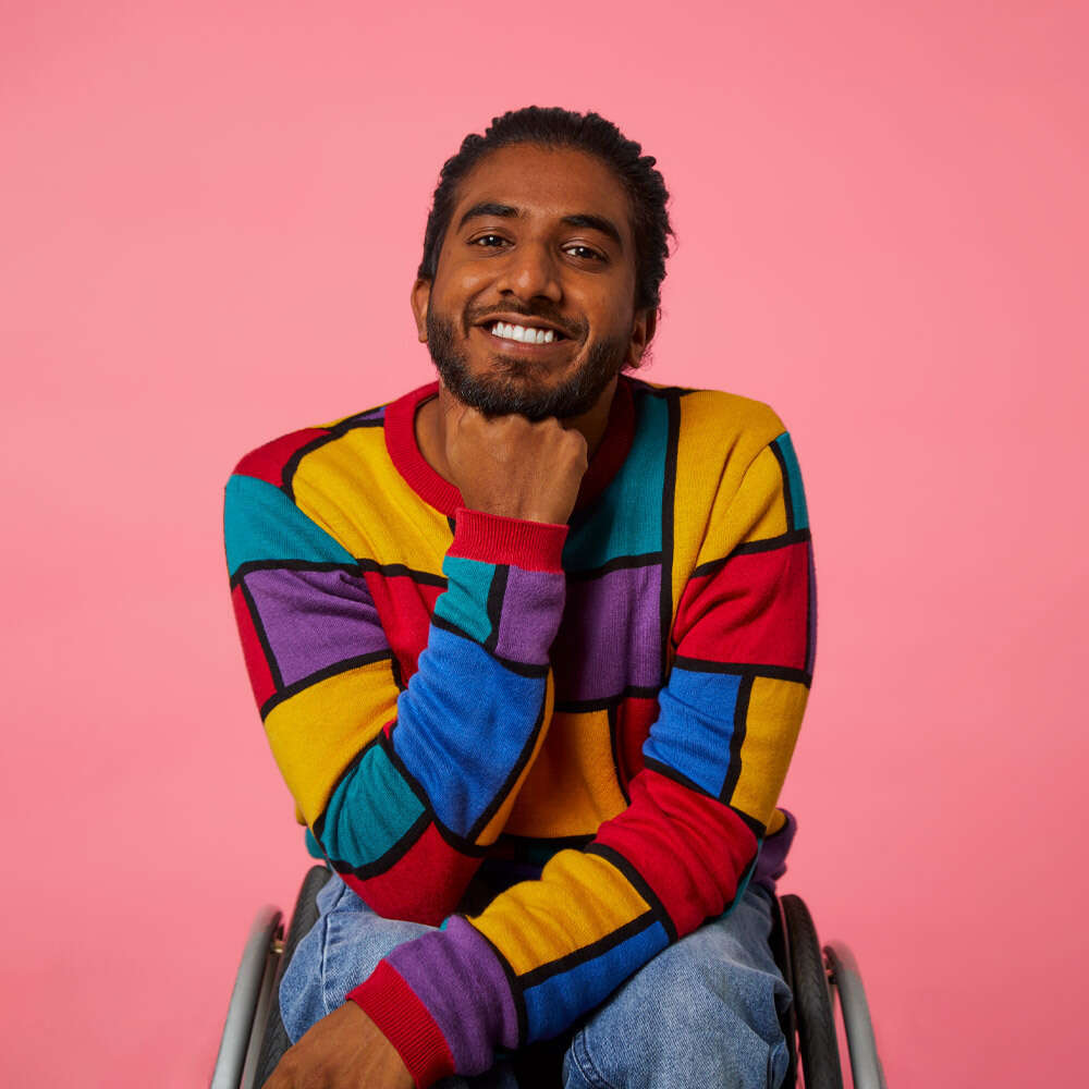 Shakti sits on his chair in front of a pink background, he wears a rainbow jumper