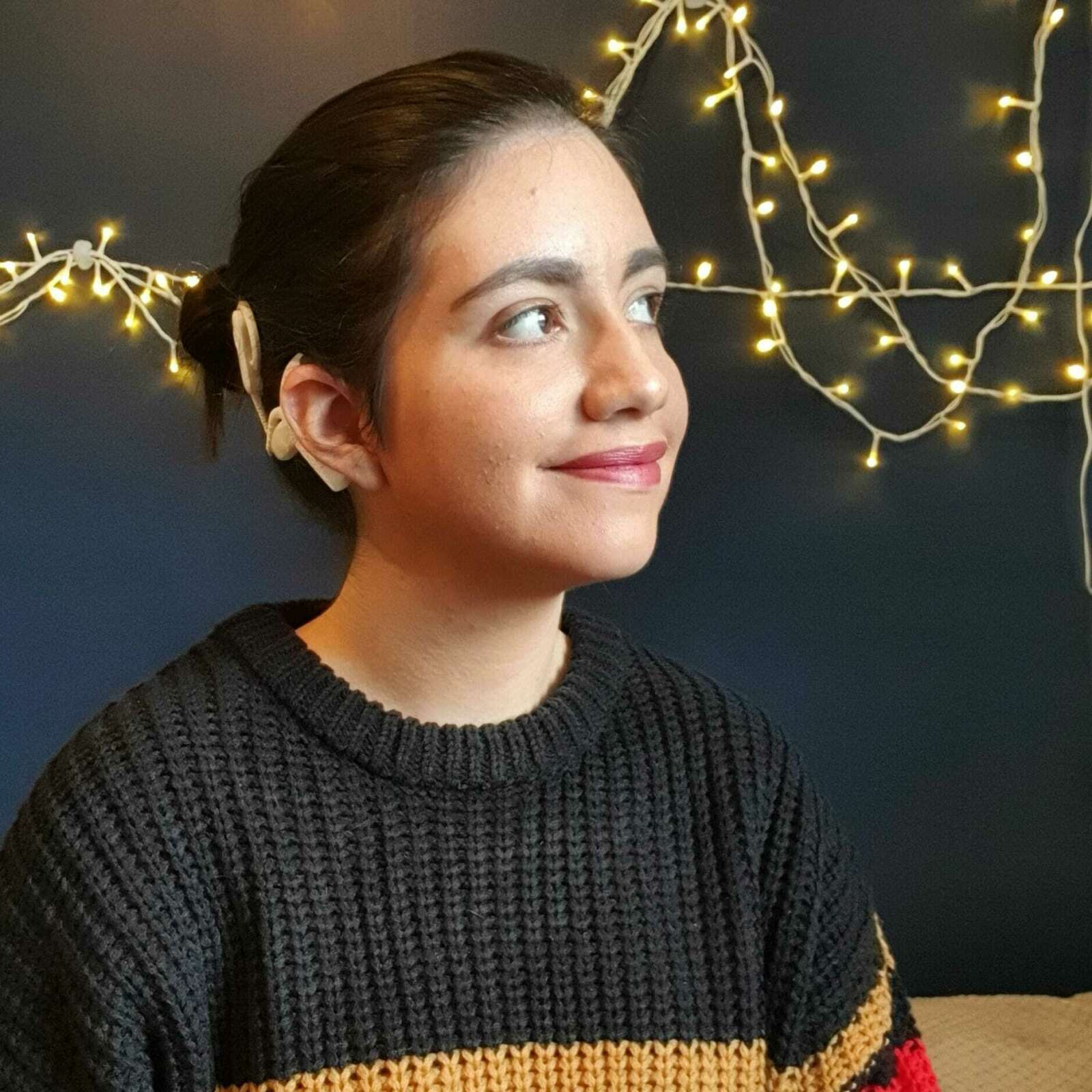 Gabriella is looking into the distance smiling. She has brown skin and dark tied black hair. She is wearing a black jumper and red lipstick.