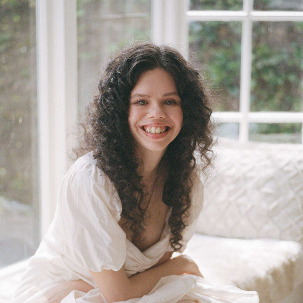 Mamie sits on her lounge day bed and wears a white dress with big curls