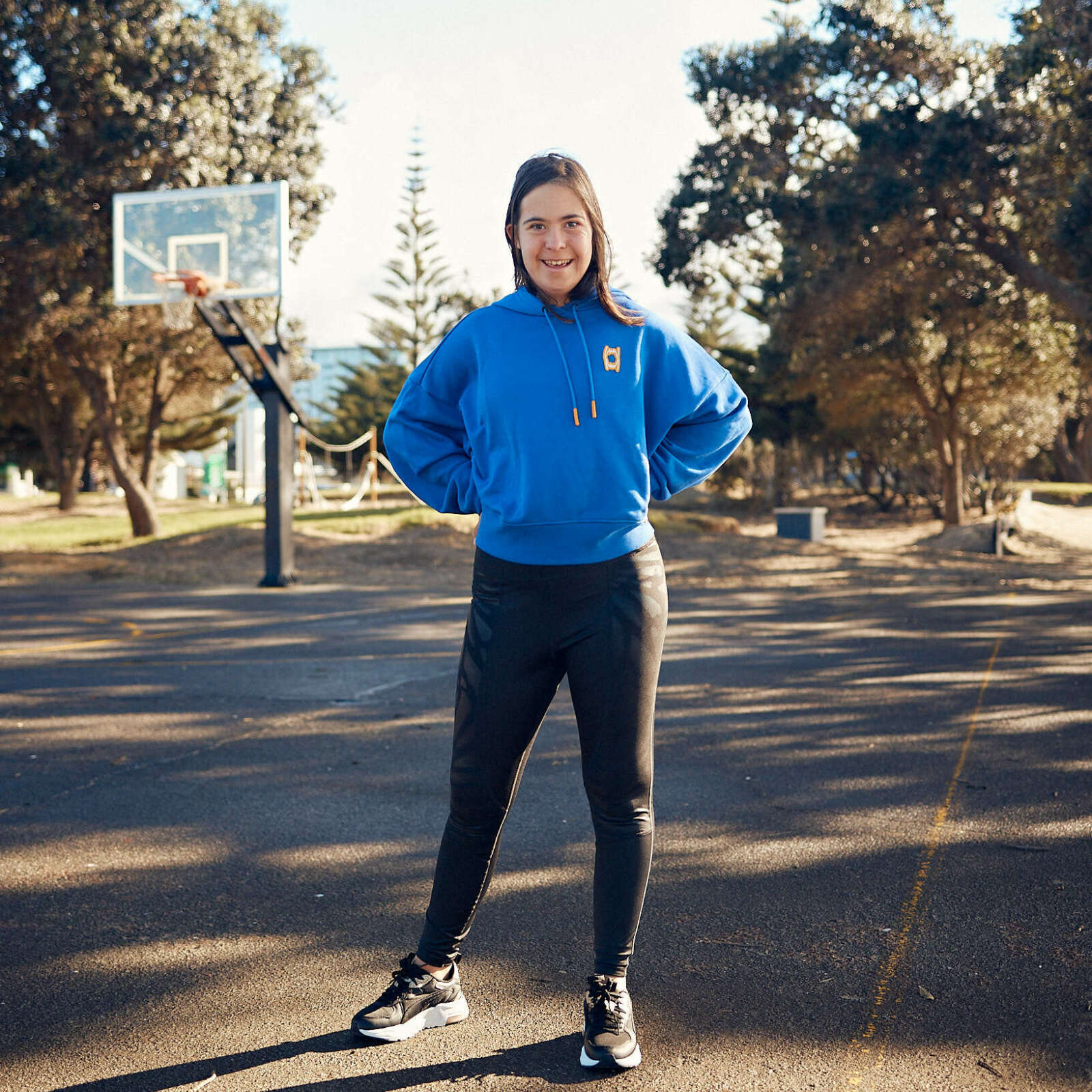 Molly is standing outside on a basketball court. She has long light brown hair and is wearing a blue hoodie with black tight leggings.