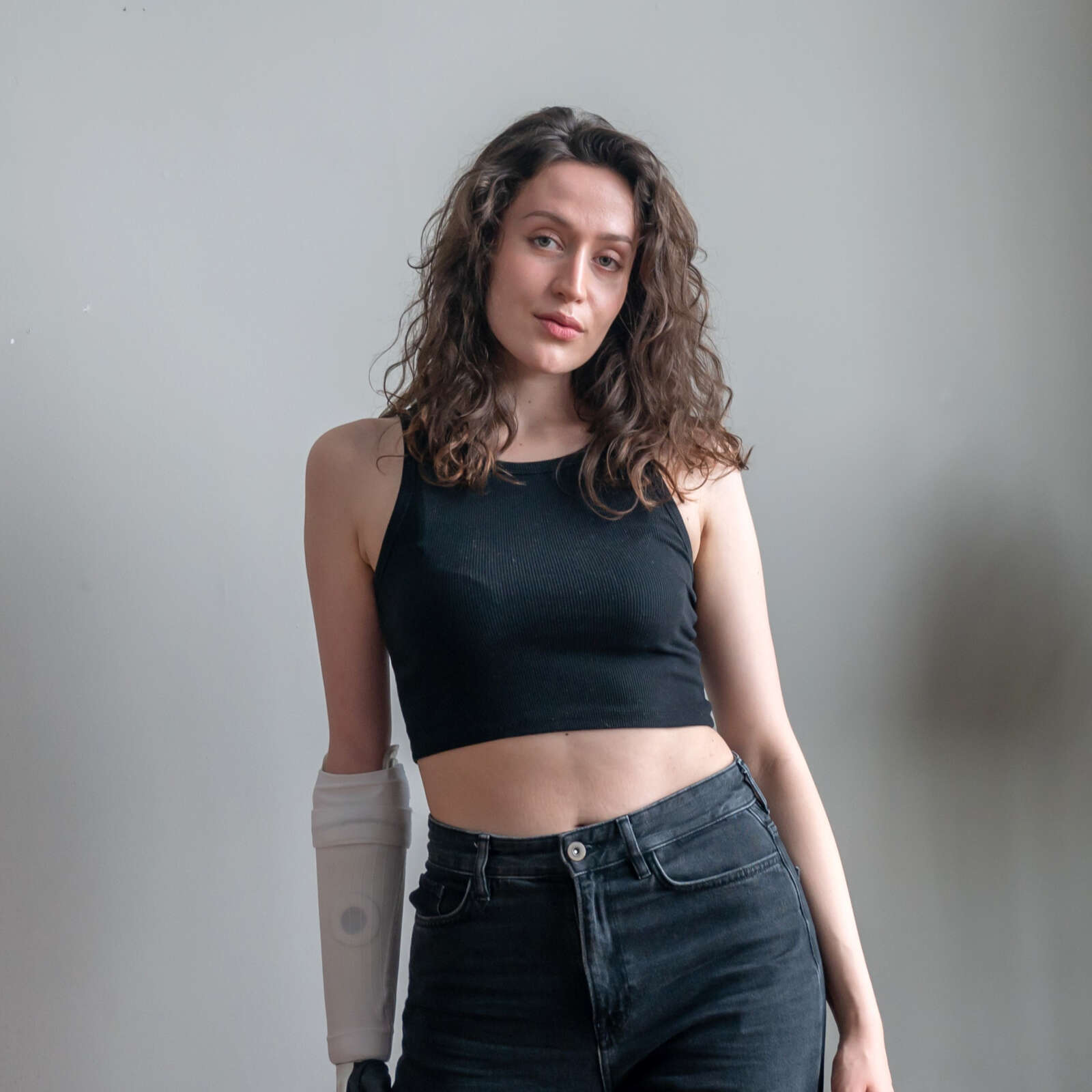 Lara is a curly-haired brunette female wearing all black. She has a white prosthetic arm and is standing against a grey backdrop.